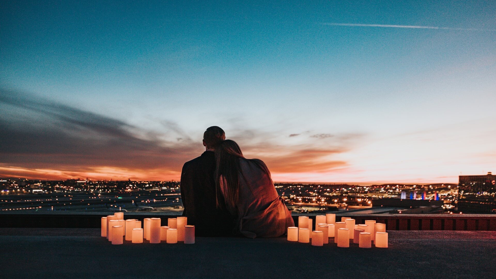 Una pareja sentada, mirando la puesta de sol sobre la ciudad, rodeada de velas